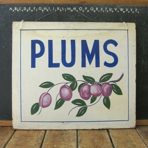 May include: A vintage square sign with the word "PLUMS" in blue lettering. Below the text is a painted branch with purple plums and green leaves. The sign has a white background and a blue border.