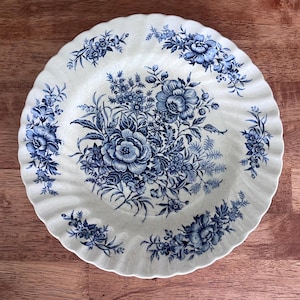 May include: A round ceramic plate with a scalloped edge, featuring a cream-colored base and a detailed blue floral pattern. The design includes a central bouquet of roses and smaller blossoms, with additional floral sprays along the rim.