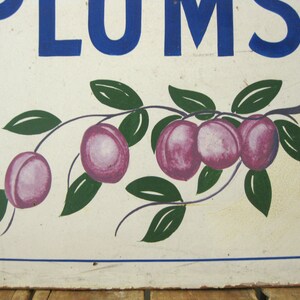May include: A vintage sign with the word "PLUMS" in blue block letters. Below, a painted branch displays several purple plums with green leaves against a white background. The sign has a rustic, aged appearance.