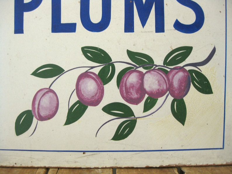 May include: A vintage sign with the word "PLUMS" in blue block letters. Below, a painted branch displays several purple plums with green leaves against a white background. The sign has a rustic, aged appearance.