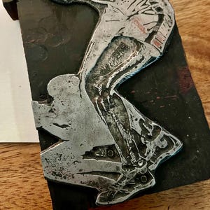 Peut inclure: Une sculpture en relief m&eacute;tallique repr&eacute;sentant une personne en mouvement, peut-&ecirc;tre en train de pratiquer un sport. La figure est rendue en argent sur un fond sombre et rectangulaire. La sculpture montre le bas du corps et les jambes de la personne.
