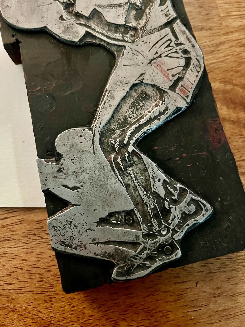Peut inclure: Une sculpture en relief m&eacute;tallique repr&eacute;sentant une personne en mouvement, peut-&ecirc;tre en train de pratiquer un sport. La figure est rendue en argent sur un fond sombre et rectangulaire. La sculpture montre le bas du corps et les jambes de la personne.