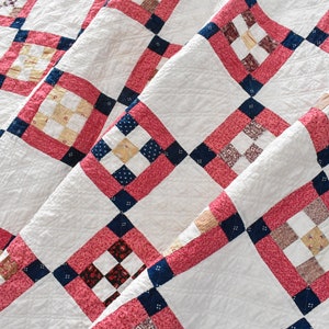 May include: A handmade quilt with a diamond pattern. The quilt features white fabric with red, navy blue, and patterned squares. The quilt is folded, showing the texture and design of the fabric.