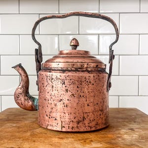 May include: Antique copper kettle with a curved spout and a large, arched handle. The kettle has a round body and a lid with a small, pointed finial. The copper has a rich, aged patina, with dark spots and variations in color, suggesting its vintage.