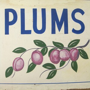 May include: A rectangular sign with the word "PLUMS" in large blue letters. Below the text is a painted illustration of a plum branch with green leaves and purple plums. The sign has a cream-colored background and a blue border.