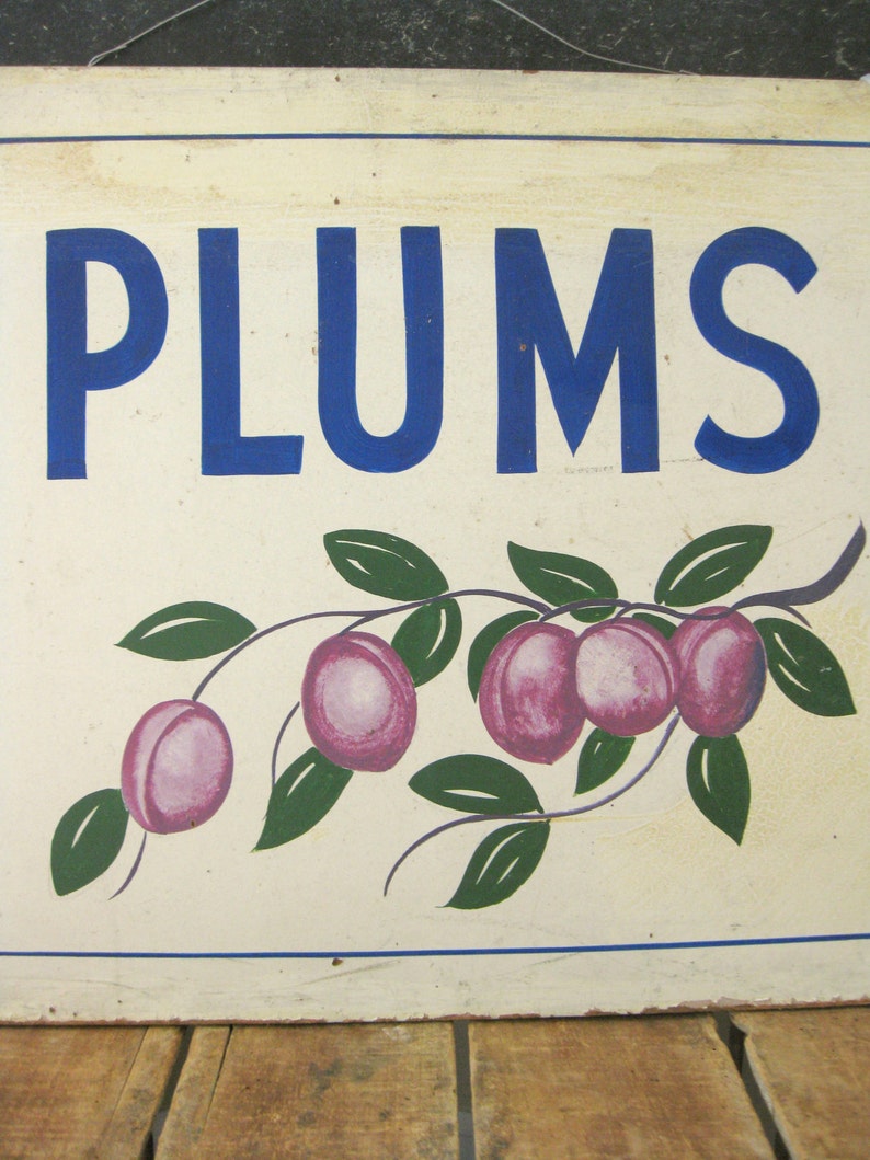 May include: A rectangular sign with the word "PLUMS" in large blue letters. Below the text is a painted illustration of a plum branch with green leaves and purple plums. The sign has a cream-colored background and a blue border.