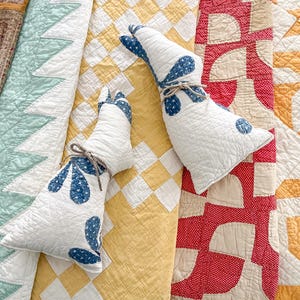 May include: Two white quilted bunny-shaped pillows with blue floral accents and tied with twine, resting on a yellow and white checkered quilt. The background includes other vintage quilts in various colors and patterns.