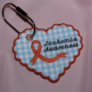 May include: A heart-shaped tag with a light blue and white gingham pattern, featuring an embroidered orange awareness ribbon and the text "Leukemia Awareness" in black script. The tag has a scalloped orange border and a silver metal keyring.