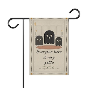 May include: A rectangular garden flag hanging on a black metal stand. The flag is beige with a border of small squares and features three black ghost illustrations, a crescent moon, candles, and the text "Everyone here is very polite" and "THE QUIET COMPANY".