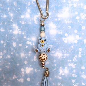 May include: A keyring featuring a silver-toned metal lobster clasp, a light blue bear bead with a smaller bear on its lap, a white bead, a brown and white spotted bead, and a light blue faux leather tassel, all connected by silver-toned spacers.