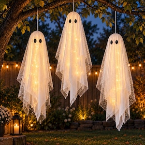 May include: Three hanging ghost decorations made of sheer white fabric with subtle glitter accents. Each ghost features two black oval eyes and is illuminated from within by warm white string lights, creating a glowing effect in the evening.