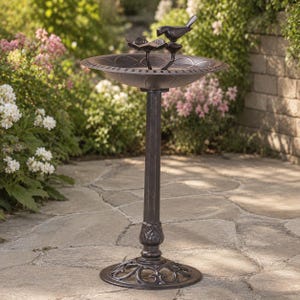 May include: A dark bronze-colored birdbath featuring a circular basin with a decorative scalloped rim. A small metal bird figurine is perched on the edge of the basin. The birdbath stands on a slender, vertical pedestal with a wide, ornate base.