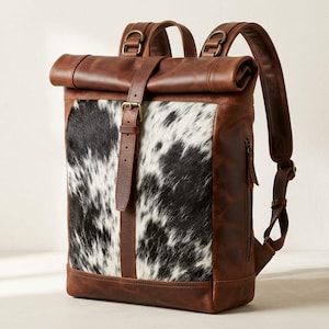 Puede incluir: Una mochila de cierre enrollable hecha de cuero marrón con un panel central que presenta un patrón de piel de vaca en blanco y negro. La bolsa incluye correas ajustables para los hombros, una correa de cuero vertical con hebilla y cremalleras laterales.