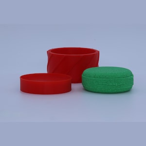 Puede incluir: Un recipiente cilíndrico rojo con textura acanalada diagonal, una tapa circular roja separada y un disco circular verde sólido con superficie texturizada. Los objetos están dispuestos juntos, mostrando el contraste de colores rojo y verde.