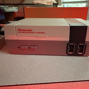 以下が含まれることがあります： 長方形のデザインのグレーとブラックのNintendo Entertainment Systemゲーム機。前面に2つのコントローラーポート、電源ボタン、リセットボタンがあり、上部には赤いNintendo Entertainment Systemのロゴがプリントされています。