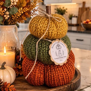 May include: A stack of three crocheted pumpkins in mustard yellow, olive green, and burnt orange. A brown stem sits on top, tied with twine. A scalloped paper tag reads Rooted Dreams Craft Co. RD Crocheted Pumpkin Stack Harvest.