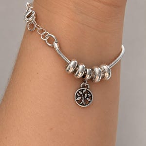 Peut inclure: Un bracelet en chaîne serpent argentée composé de quatre perles lisses et arrondies ainsi qu'un pendentif circulaire représentant un arbre avec ses branches et ses feuilles. Le bracelet est doté d'un fermoir mousqueton et d'une chaînette réglable.