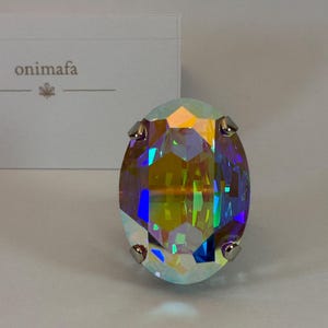 Peut inclure: Une bague en cristal irisé de forme ovale avec une surface à facettes multiples reflétant des teintes jaunes, bleues et violettes. Le cristal est fixé par quatre griffes argentées. En arrière-plan, une carte blanche affiche le texte onimafa.
