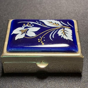 May include: A rectangular metal pill box with a hinged lid featuring a glossy deep blue enamel inlay. The lid is decorated with a hand-painted white floral motif accented by delicate gold-coloured dots and thin gold-coloured lines.