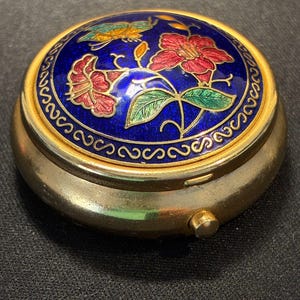 Op de afbeelding: Een ronde goudkleurige metalen pillendoos met een blauw geëmailleerd deksel versierd met rode bloemen, groene bladeren en een kleine vlinder. Het deksel is omrand door een goudkleurige metalen rand met een herhalend krulpatroon en een drukknop.