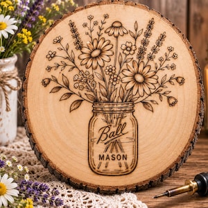 May include: A circular wood slice featuring a pyrography design of a bouquet of wildflowers in a Ball Mason jar. The wood has a natural bark edge, and the detailed wood-burned illustration depicts daisies, lavender, and various small floral stems.
