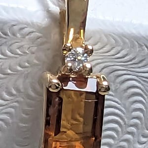 Op de afbeelding: Een gouden hanger met een rechthoekige, amberkleurige edelsteen. Boven de hoofdsteen bevindt zich een kleine cluster van heldere stenen. De hanger hangt aan een witte ketting.