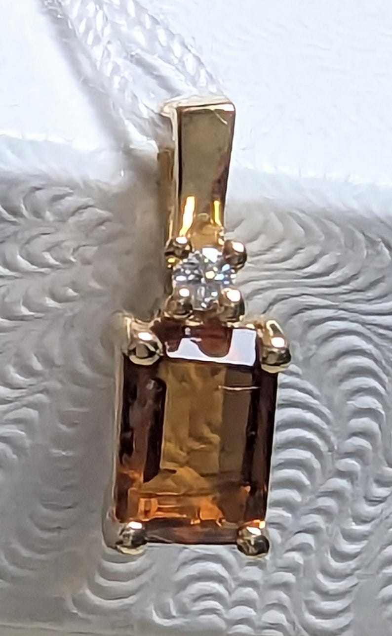 Op de afbeelding: Een gouden hanger met een rechthoekige, amberkleurige edelsteen. Boven de hoofdsteen bevindt zich een kleine cluster van heldere stenen. De hanger hangt aan een witte ketting.