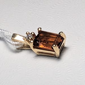 Op de afbeelding: Een gouden hanger met een rechthoekige, bruine edelsteen. Een kleine, heldere steen is naast de edelsteen gezet. De hanger is bevestigd aan een wit koord. De zetting is van goud.