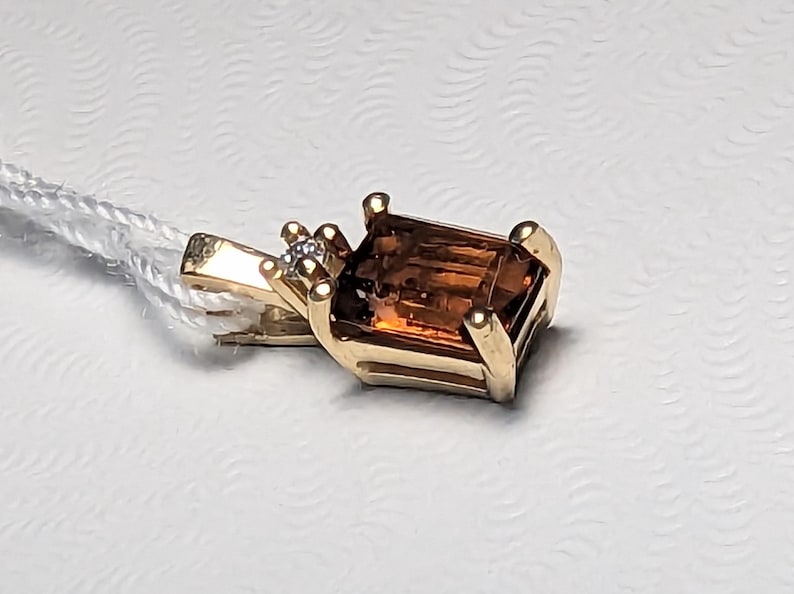 Op de afbeelding: Een gouden hanger met een rechthoekige, bruine edelsteen. Een kleine, heldere steen is naast de edelsteen gezet. De hanger is bevestigd aan een wit koord. De zetting is van goud.