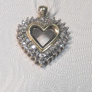 Op de afbeelding: Een gouden hartvormige hanger, omringd door kleine, sprankelende stenen. De hanger heeft een hartvormige uitsparing in het midden en hangt aan een dun, wit koord. Waarschijnlijk gemaakt van goud en diamanten.