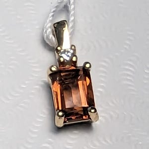 Op de afbeelding: Een gouden hanger met een rechthoekige, oranjebruine edelsteen. De hanger heeft een kleine diamant en vier gouden klauwen. Hij hangt aan een wit touwtje.
