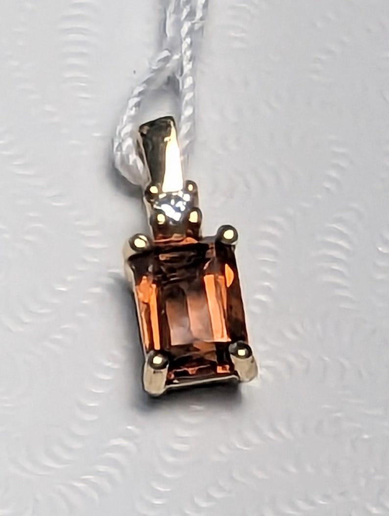 Op de afbeelding: Een gouden hanger met een rechthoekige, oranjebruine edelsteen. De hanger heeft een kleine diamant en vier gouden klauwen. Hij hangt aan een wit touwtje.