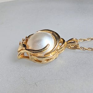 Op de afbeelding: Een gouden ketting met een hanger met een grote witte parel in een decoratief gouden frame. De ketting is een fijne gouden kabelketting.