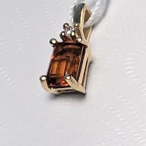 Op de afbeelding: Een gouden hanger met een rechthoekige, amberkleurige edelsteen. De hanger heeft een kleine diamant en hangt aan een wit koord. De zetting van de hanger heeft een klassiek ontwerp. De edelsteen is ongeveer 1,5 cm lang.