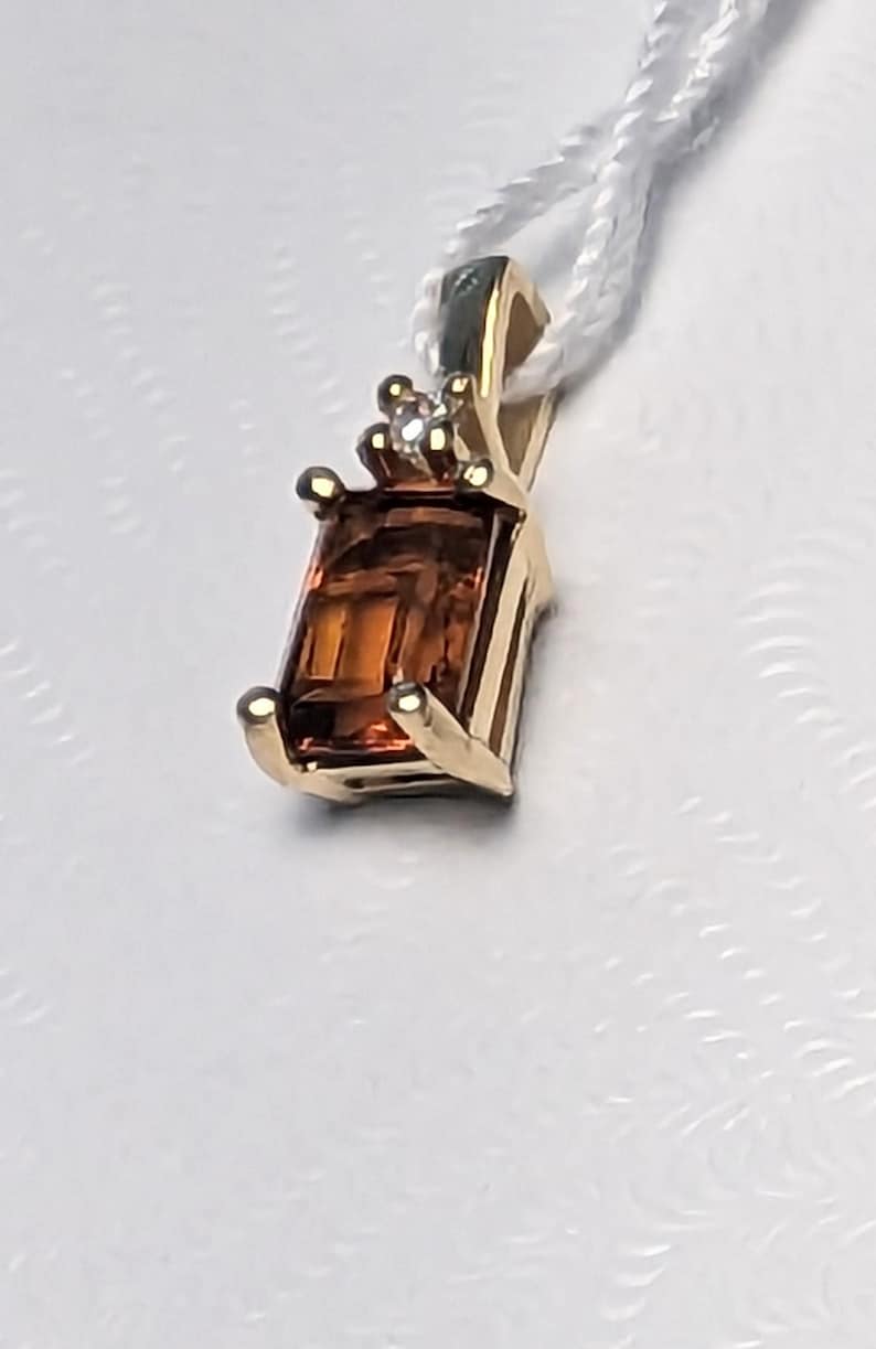 Op de afbeelding: Een gouden hanger met een rechthoekige, amberkleurige edelsteen. De hanger heeft een kleine diamant en hangt aan een wit koord. De zetting van de hanger heeft een klassiek ontwerp. De edelsteen is ongeveer 1,5 cm lang.