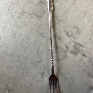 May include: A silver-toned metal cocktail fork featuring a long, twisted handle and a decorative, beaded-edge top section. The fork has three slender tines and a polished finish, showcasing a slender, elongated design for serving small appetizers.