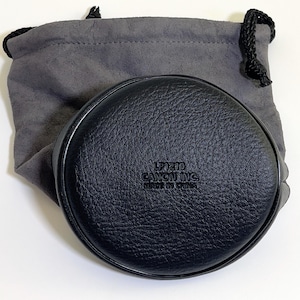 Peut inclure: Un étui à objectif circulaire noir avec une surface texturée et le texte en relief LP1219 CANON INC. MADE IN CHINA. L'étui est placé devant une pochette à cordon grise en matière douce semblable à du daim avec des fermetures noires.