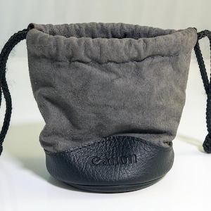 Peut inclure: Une pochette à cordon en daim gris avec une base en cuir noir portant le logo Canon en relief. La pochette est dotée de deux cordons tressés noirs de chaque côté pour fermer le haut. Le design est simple et fonctionnel pour le rangement.
