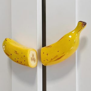 May include: A pair of yellow banana-shaped cabinet knobs mounted on white doors. The left knob is a cross-section showing a light-colored interior with brown seeds, while the right knob shows the curved stem end of the banana with brown spots.