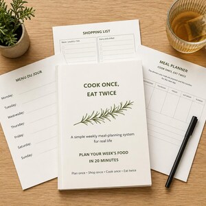 May include: A white meal planning booklet titled Cook Once, Eat Twice with a rosemary sprig illustration, accompanied by a Menu Du Jour sheet, a Shopping List, and a Meal Planner sheet. The set includes a black pen and a small potted green plant.