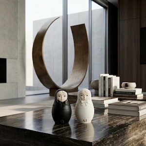 May include: Two nesting dolls, one in matte black and one in matte white, each featuring a sculpted face, a head covering, and a small bow at the neck. Each doll is marked with a small letter M at the base, showcasing a smooth, rounded form.