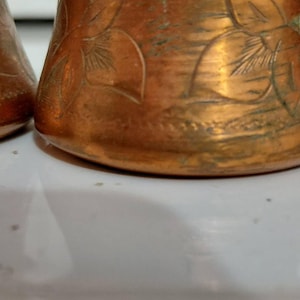 May include: A copper vessel featuring a tapered body with an engraved floral pattern and a metallic handle attached to the side. The surface displays a warm, reddish-brown metallic finish with visible signs of oxidation and fine etched details.