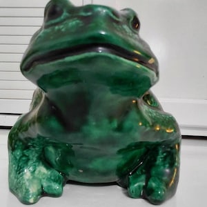 May include: A ceramic frog figurine with a glossy finish, featuring a deep emerald green glaze with darker blackish-green mottled patches. The frog is depicted in a seated, forward-facing position with a wide mouth and textured skin.