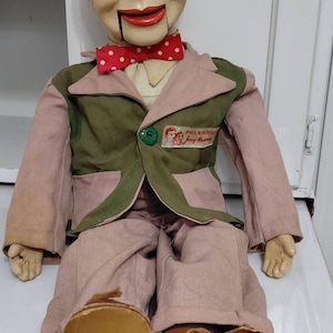 May include: A ventriloquist dummy with reddish-brown hair, blue eyeshadow, and a red mouth. It wears a pinkish-tan suit with a green vest, a red and white polka-dot bow tie, and brown shoes. A label on the vest reads Paul Winchell's Jerry Mahoney.