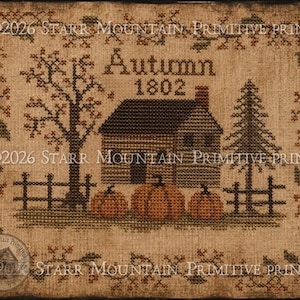 May include: A cross-stitch pattern on tan fabric featuring a house, three orange pumpkins, a tree, and a pine tree. The text reads "Autumn 1802" and "©2026 STARR MOUNTAIN PRIMITIVE PRINTS". A decorative border of vines and leaves surrounds the central scene.