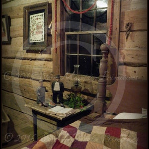 May include: A rustic log cabin bedroom decorated for Christmas. A red and white quilt covers the bed, and a small table with a book and a few decorations sits beside it. Two small wooden figures stand on the table. The room has a wooden floor and walls, and a window with a red garland hanging from it.