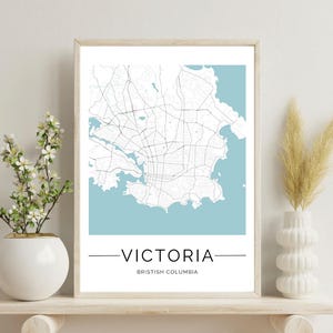 May include: A framed map print of Victoria, British Columbia. The map is in black and white with a light blue background. The city name is printed in black below the map.