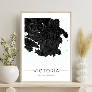 May include: Framed black and white map print of Victoria, British Columbia. The city map is detailed with streets and waterways. The word "VICTORIA" is centered below the map, with "BRITISH COLUMBIA" underneath. The print is displayed on a wooden shelf with decorative vases.