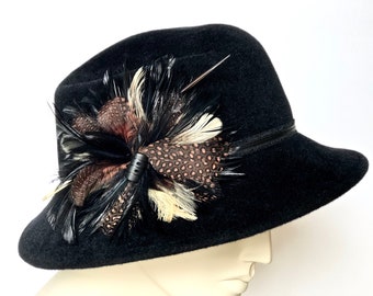 May include: A black fedora hat with a decorative arrangement of feathers in shades of brown, black, and cream. The hat features a black band and a silver pin. The feathers are clustered on the side of the hat, adding a touch of elegance.
