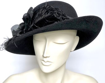 May include: A black hat with a wide brim, adorned with black feathers and fabric flowers. The hat is displayed on a mannequin head. The hat's design is classic and elegant, suitable for special occasions.